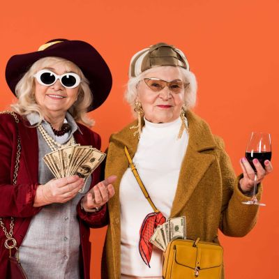 cheerful senior woman holding money near friend with glass of wine in hand isolated on orange